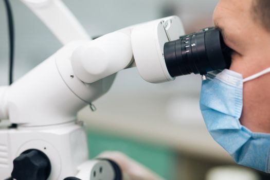 Dentist using dental microscope and examining woman's teeth. Medium shot of male dentist in scrubs and face mask using dental microscope while examining patient.