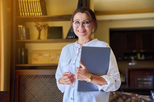 Portrait of mature business woman in glasses with laptop in her hands, living room interior or office. Successful confident smiling 40s female looking at camera. Business, people, management, staff