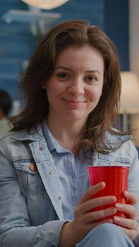 Portrait of woman sitting on sofa similing at camera while drinking beer late at night during holiday party. In background multi-ethnic friends gathering together for celebrating birthday