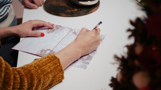 Close up of woman hand signing christmas gift card for holiday celebration. Adult writing on postcard paper for seasonal festivity, giving presents to relatives. Couple preparing for christmas.