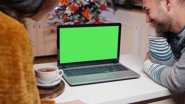 Festive couple looking at horizontal green screen on christmas celebration. People watching chroma key with isolated background and mockup template on laptop for holiday festivity.