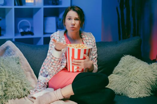 Woman sitting in living room sofa couch watching interesting program and pointing sharing with eating popcorn at night