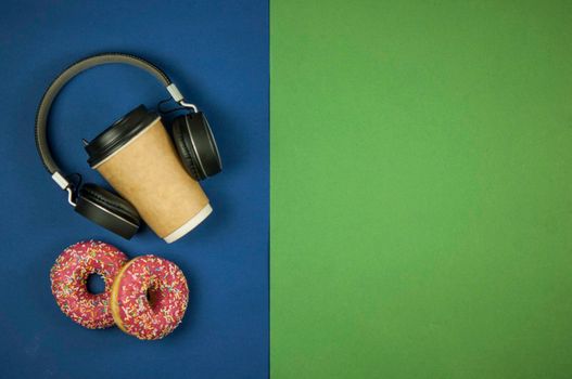 A paper coffee cup with two doughnuts and wireless headphones lie on a green and blue background. A place to copy. Top view. Audiobook concept. Distance learning. Close-up, copy the space, the background