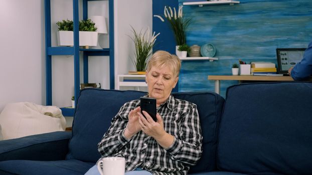 Overjoyed middle-aged woman sitting on couch in living room talking on video call on smartphone gadget, happy excited senior female greeting speak on web using smartphone, technology concept