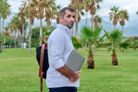 Portrait of serious confident mature male with laptop outdoors. Businessman with backpack, laptop in hands, tropical park nature. Business, technology, freelance, lifestyle, middle age, business trip