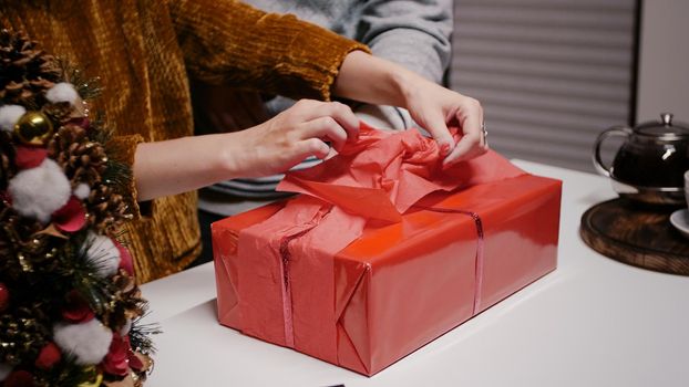 Close up of hand decorating present box with wrapping paper and bow with ribbon. Festive woman preparing gift for family, using ornaments for christmas eve celebration. Winter festivity