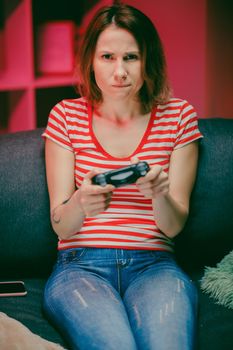 Determined girl playing a video game at living room at night. Gamer woman sitting on a sofa, playing in video games on a console and using a wireless controller.