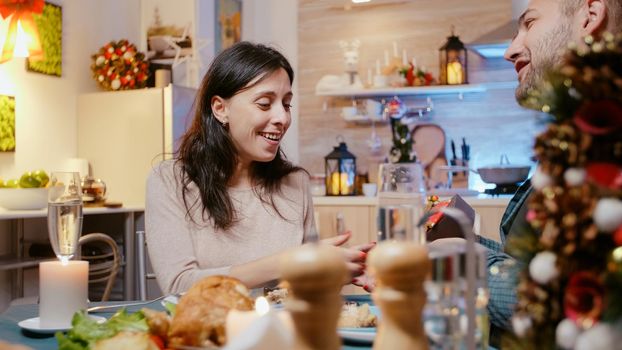 Man giving present box to woman for christmas celebration while enjoying festive dinner. Adult receiving gift box decorated with ribbon and bow tie from partner, eating meal and drinking