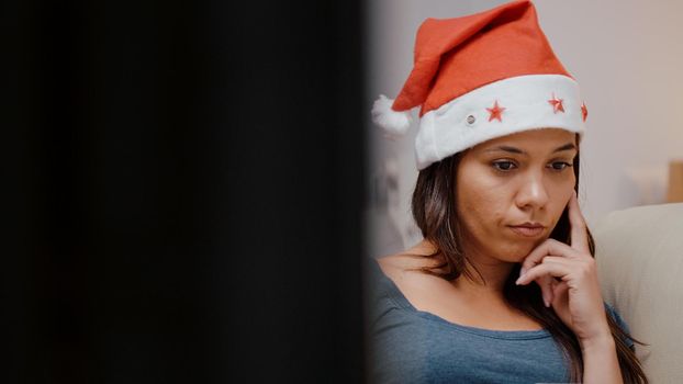 Close up of unhappy person working on laptop at television. Festive woman wearing santa hat using modern device on christmas eve. Adult laying on couch with gadget on holiday.