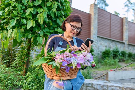 Middle-aged woman in garden with basket of fresh cut spring flowers using smartphone. Rest, gardening, lifestyle, leisure, emotions, spring, nature, technologies, middle age concept