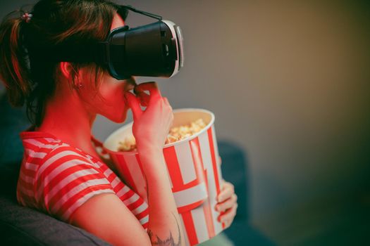 Young woman wear VR headset and watch movie with popcorn at night. American woman sitting on the sofa in the VR glasses and watching something while eating popcorn. Indoor