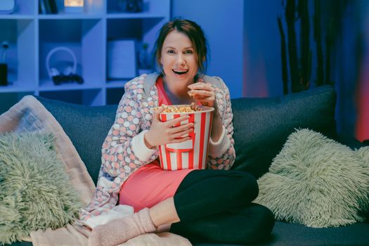Happy woman sitting in living room sofa couch watching funny interesting program and pointing sharing with eating popcorn at night