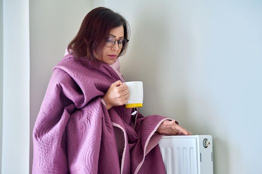 Heating during the cold winter autumn season. An adult woman warming herself with a blanket, a central heating radiator, hot tea.