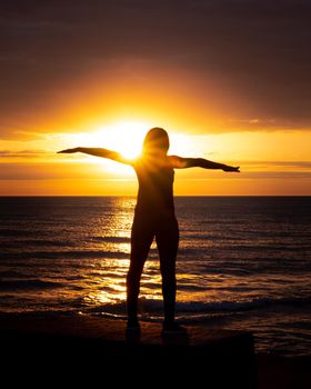 A beautiful woman's silhouette with her arms outstretched standing in front of Lake Michigan water reflecting an incredible sunrise with bright orange and yellow colors.