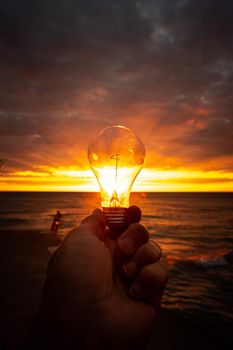 A man holds out a clear lightbulb in front of a colorful sunrise over Lake Michigan with rays of yellow, orange and pink filling the glass of the bulb making a beautiful background image.