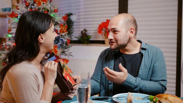 Man receiving gift box with ribbon from woman while eating chicken at festive dinner on christmas eve. Partners celebrating seasonal holiday with presents and meal with champagne