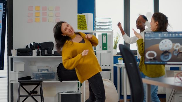 Happy diverse businesswomen dancing in company office celebrating success of project, singing and having fun during working hours. Excited multiethnic engineers using computer showing cad software