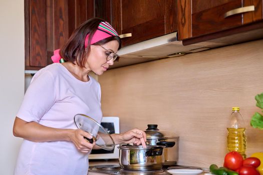 Middle-aged woman housewife preparing food in a saucepan on the hob in the home kitchen. Cooking, homemade food, lifestyle, household concept