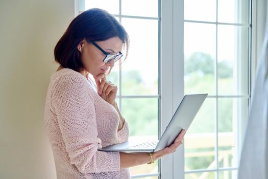 Mature businesswoman in glasses with laptop in her hands near window. Remote work, education, business, freelance, home office, technology concept