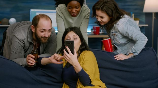 Woman showing comedy video on phone to her multiracial friends socializing together during night party. Group of mixed race people sitting on sofa, drinking beer, chilling in living room