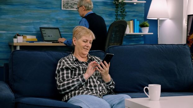 Happy middle aged mature woman enjoying using mobile apps texting typing messages sit on sofa, smiling old adult lady holding smartphone looking at cellphone screen browsing social media at home