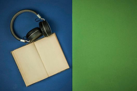 Black headphones with old open book with blank pages is lying on green and blue background. Copy space. Top view. Audio book concept. Distance learning. Close up, background. 