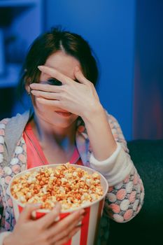 Portrait shot of the woman with popcorn sitting on the sofa watching something scary while eating popcorn and being afraid. Inside