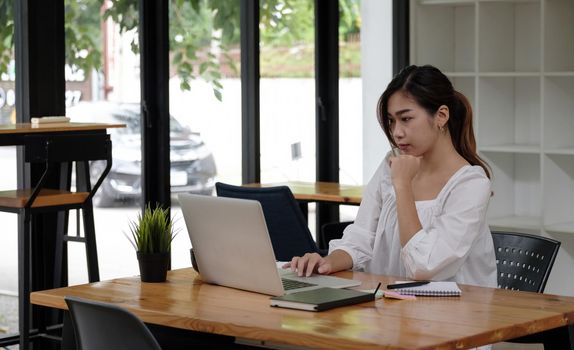 Focused asian business woman working studying online in office looking at laptop, serious employee or student watching webinar writing information in notebook sitting at desk.