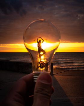 A man holds up a lightbulb in front of a woman's ballerina silhouette on the beach at sunrise with majestic orange and yellow clouds reflecting on the water.