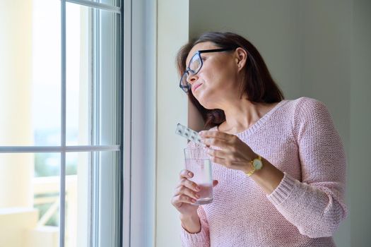 Middle-aged woman with pills and glass of water in her hands, at home near window, copy space. Mature women's health, pharmacology, vitamins, supplements, therapy, treatment