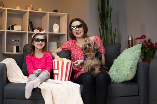 Caucasian woman and her little daughter in 3D glasses sitting on the sofa and watching comedy film on TV while eating popcorn and laughing. Indoors.