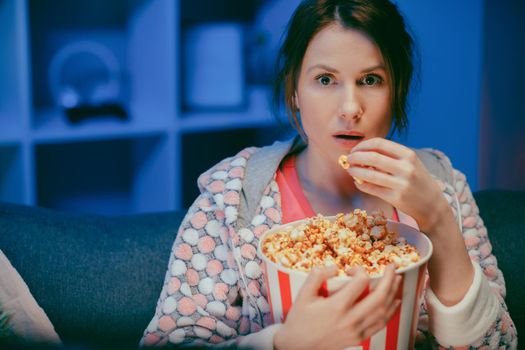 Portrait shot of the woman with popcorn sitting on the sofa watching something scary while eating popcorn and being afraid. Inside