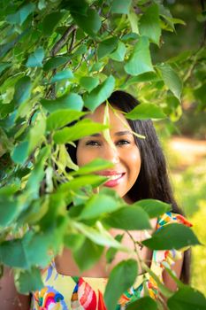 A beautiful young mixed race African American woman smiles with her teeth from behind a tree branch with leaves obscuring her face and upper body as she wears a yellow floral print dress.