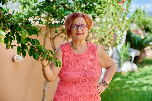 Outdoor portrait of a beautiful elderly 70s woman in the garden. Retirement age female with glasses looking at camera, near tree. Beauty, age, health, pensioner, retired older people concept