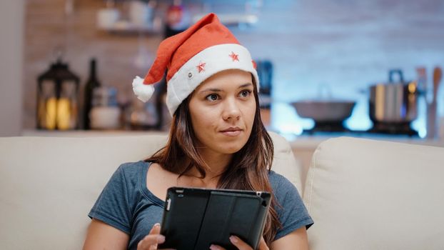 Person wearing santa hat and working on digital tablet. Woman holding gadget and watching television on christmas eve holiday. Festive adult with device and technology in winter season