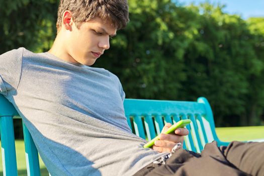 Serious guy teenager using smartphone, having rest, sitting on bench in park. Youth, technology, lifestyle, adolescence concept