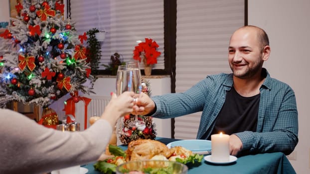 Close up of man enjoying festive dinner and clinking glasses of champagne with woman on christmas eve. Couple celebrating winter holiday with traditional food and alcohol on table.