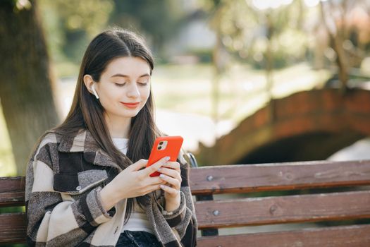 Attractive woman browsing on mobile phone in public park. City, urban background. Happy girl listening to music in the park using mobile phone and wearing headphones.