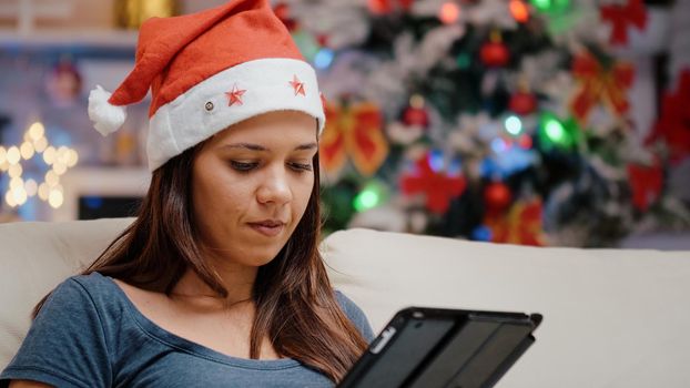 Close up of woman with santa hat holding digital tablet and watching television on christmas eve holiday. Festive adult working with device while looking at movie on TV for celebration