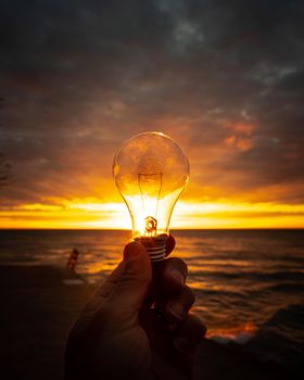 A man holds out a clear lightbulb in front of a colorful sunrise over Lake Michigan with rays of yellow, orange and pink filling the glass of the bulb making a beautiful background image.