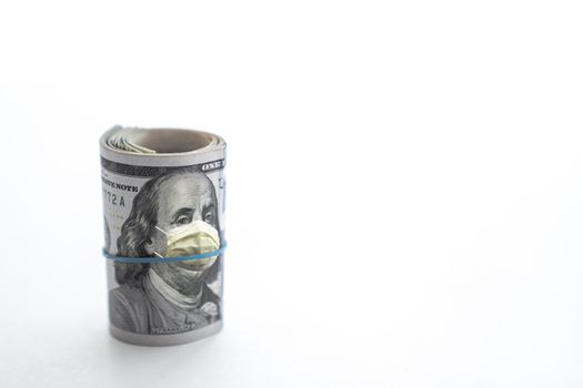 Rolled up wad of cash with hundred dollar bill on top and blue rubber band around the face of Benjamin Franklin wearing a yellow COVID-19 protective face covering or mask isolated on white background.
