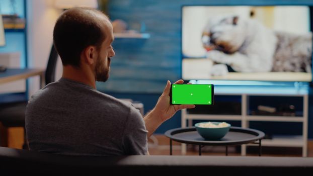 Young man watching horizontal green screen on smartphone display sitting at home. Copy space equipment with chroma key and isolated background media for mockup template and design
