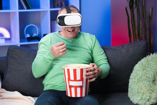 Senior Caucasian Man With a Box of Popcorn in His Hand Watching Video Using Virtual Reality Headset Sitting on Sofa Eating Popcorn in Room
