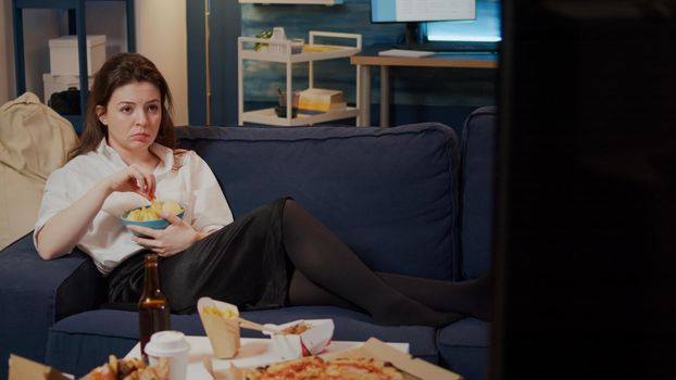Young woman laying on couch holding bowl of chips watching television at home. Person with takeaway fast food on table relaxing on sofa after work while having snack in living room