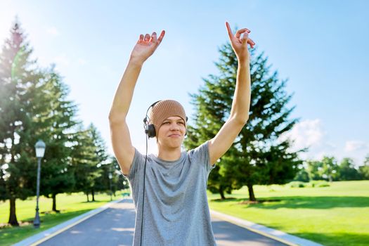 Portrait of cheerful hipster guy in knitted hat with headphones, dancing, hands up. Smiling teenager listening to music, summer in park. Hobbies, leisure, creativity, youth, lifestyle and technology