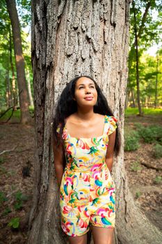 Beautiful portrait of a happy mixed race African American woman wearing a yellow floral dress leaning against a large rough bark covered tree trunk in a forest and looks up into the distance.