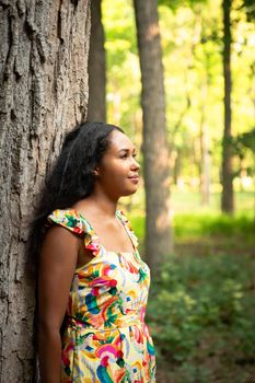 Beautiful portrait of a happy mixed race African American woman wearing a yellow floral dress leaning against a large rough bark covered tree trunk in a forest and looks off into the distance.