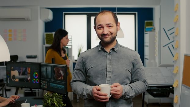 Portrait of videographer standing in start up creative agency office holding cup of coffee looking at camera and smiling. Man working in multimedia studio production editing video in modern workplace.