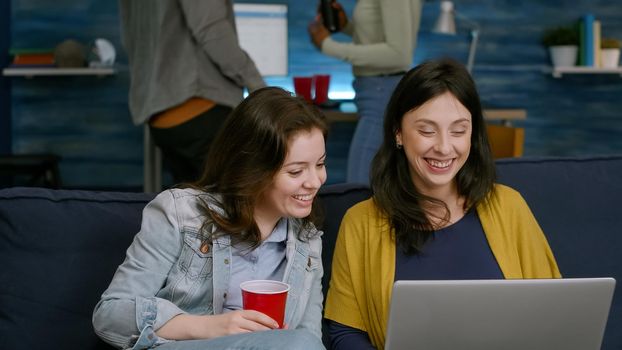 Excited friends watching entertainment show on laptop while sitting on couch in living room late at night during party. African woman with friend man drinking beer, dancing together enjoying free time