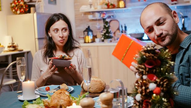 Man and woman exchanging presents at festive dinner, celebrating christmas eve. Cheerful people giving gift boxes with wrapping paper and ribbon while enjoying chicken meal and drinks